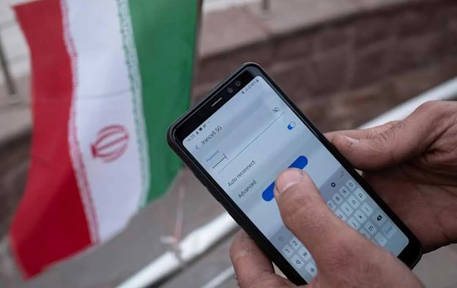 کەسێک وشەی نهێنیی ئینتەرنێت لەسەر مۆبایلەکەی دەنووسێت و لەدواوە ئاڵای ئێران دەرکەوتووە